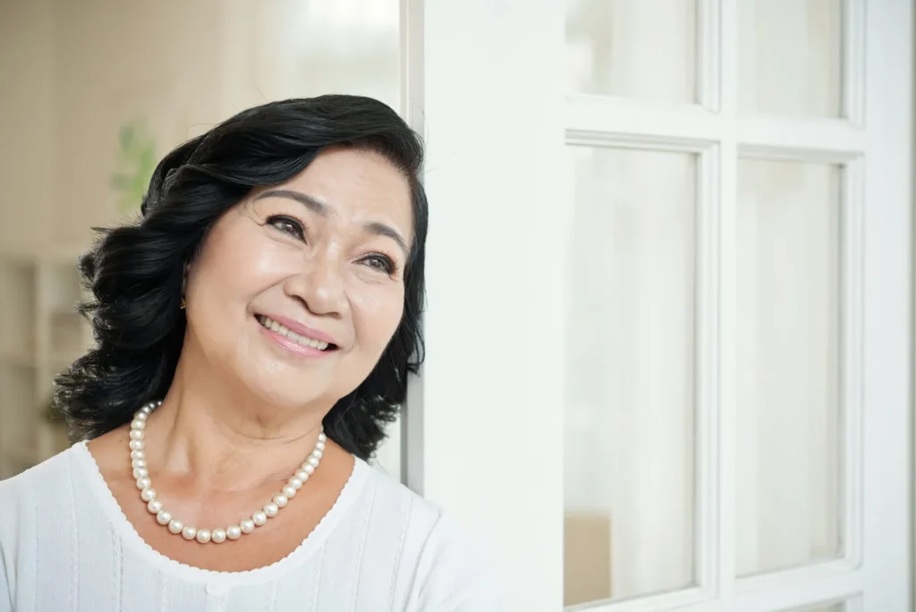 smiling senior asian woman leaning door home looking away